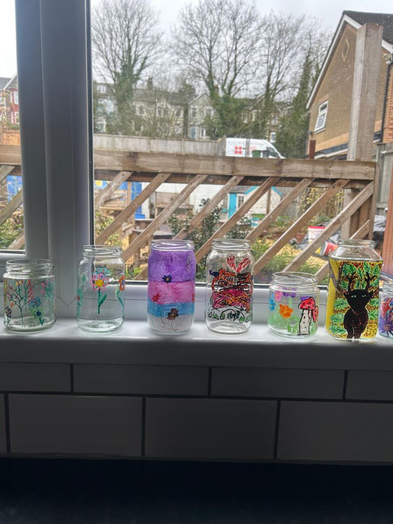 A row of glass jars hand-painted with flowers, a butterfly and a cat under a tree, lined up on a windowsill
